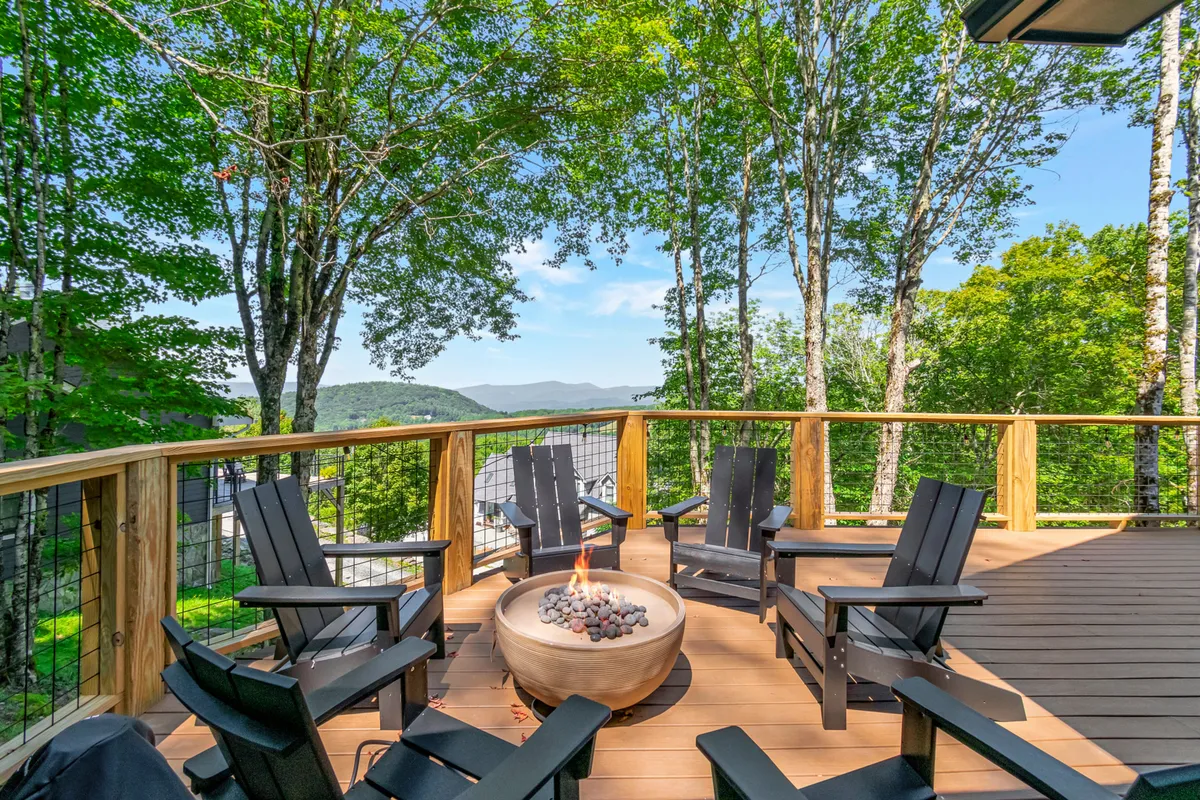 A spacious wooden deck with Adirondack chairs surrounding a fire pit, with a wooden railing framing a gorgeous mountain view. Lush greenery surrounds the deck, and the fire pit is ready for gathering. The inviting setup is perfect for intimate gatherings or quiet moments, and the mountain landscape in the background reinforces the scenic, outdoor vibe.