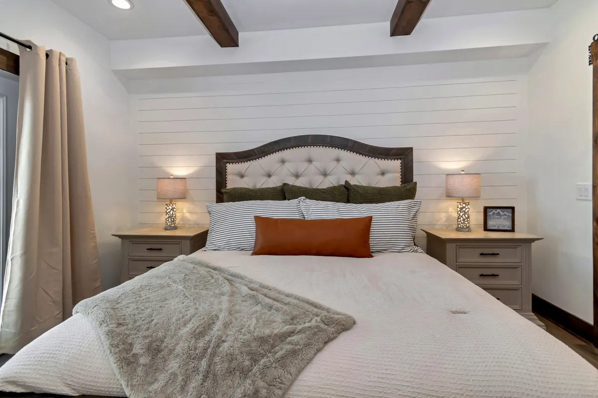 A serene bedroom features a tufted headboard, striped bedding, decorative pillows, and matching nightstands with soft-glowing lamps. The space is styled for comfort and relaxation, hinting at luxurious linens and offering glimpses of natural light through windows.