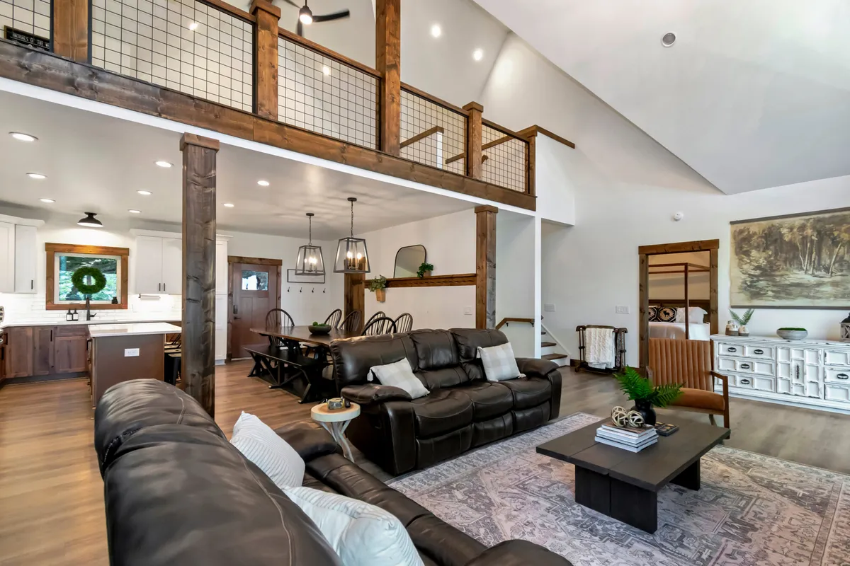 This image captures a welcoming open-concept living space with leather sofas gathered around a coffee table, an open kitchen, and a dining area in view. Large windows bring in abundant natural light while offering a glimpse of outdoor scenery. Decorative wall art and cozy furnishings create a sense of warmth and style.