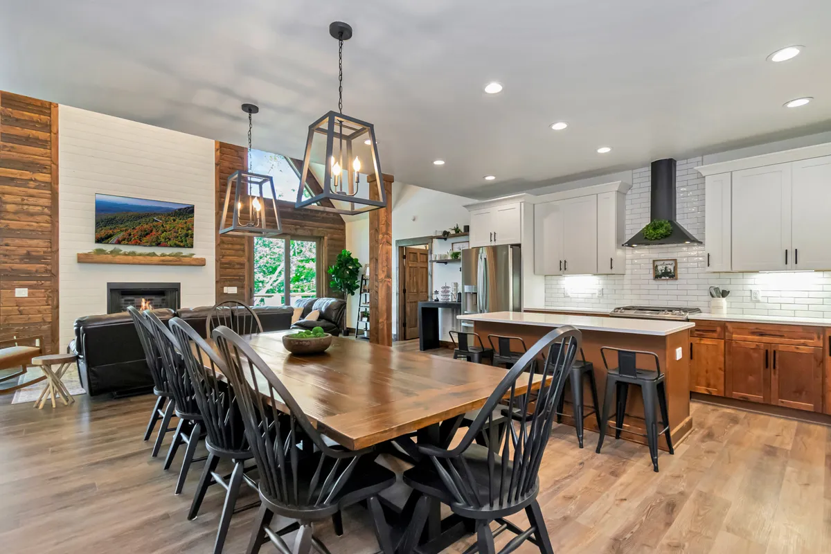 A wide shot reveals a spacious wooden dining table as the centerpiece, with a kitchen island and the living area—including a fireplace—visible in the background. Artwork and decorative touches add personality, while large windows flood the space with sunlight, framing outdoor views.