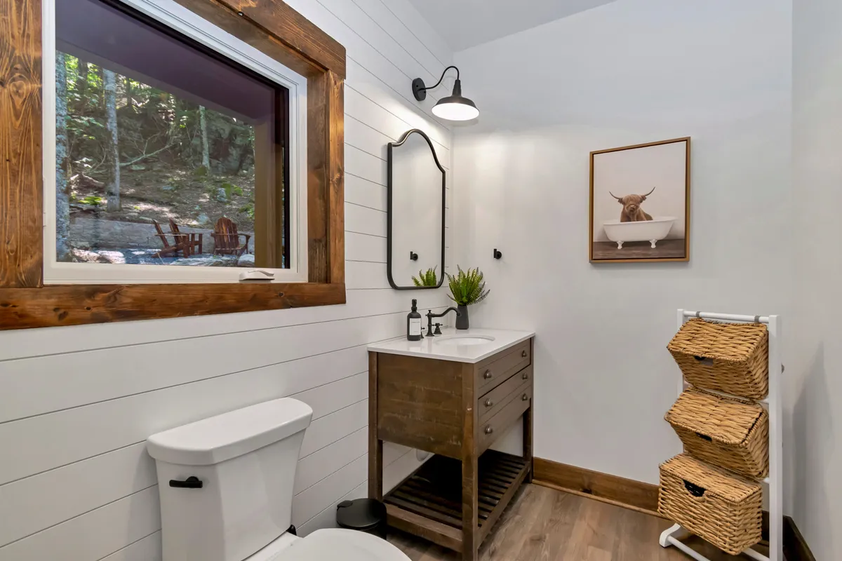 This image features a window with a view of outdoor seating, a wooden vanity, a decorative mirror, and basket storage. The window provides a clear connection to the outside world and hints at the inviting natural setting just beyond the cabin. The scene feels calm and comfortable, with a subtle suggestion of fresh air and the opportunity to relax after outdoor adventures.