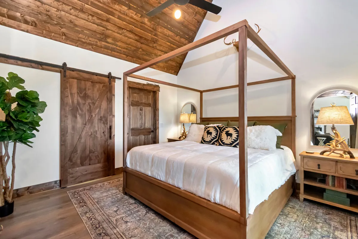 A tranquil bedroom with a four-poster bed, wooden ceiling, sliding barn door, decorative plants, and nightstands with lamps. The room exudes rustic charm and an inviting, restful vibe, enhanced by soft, natural lighting.