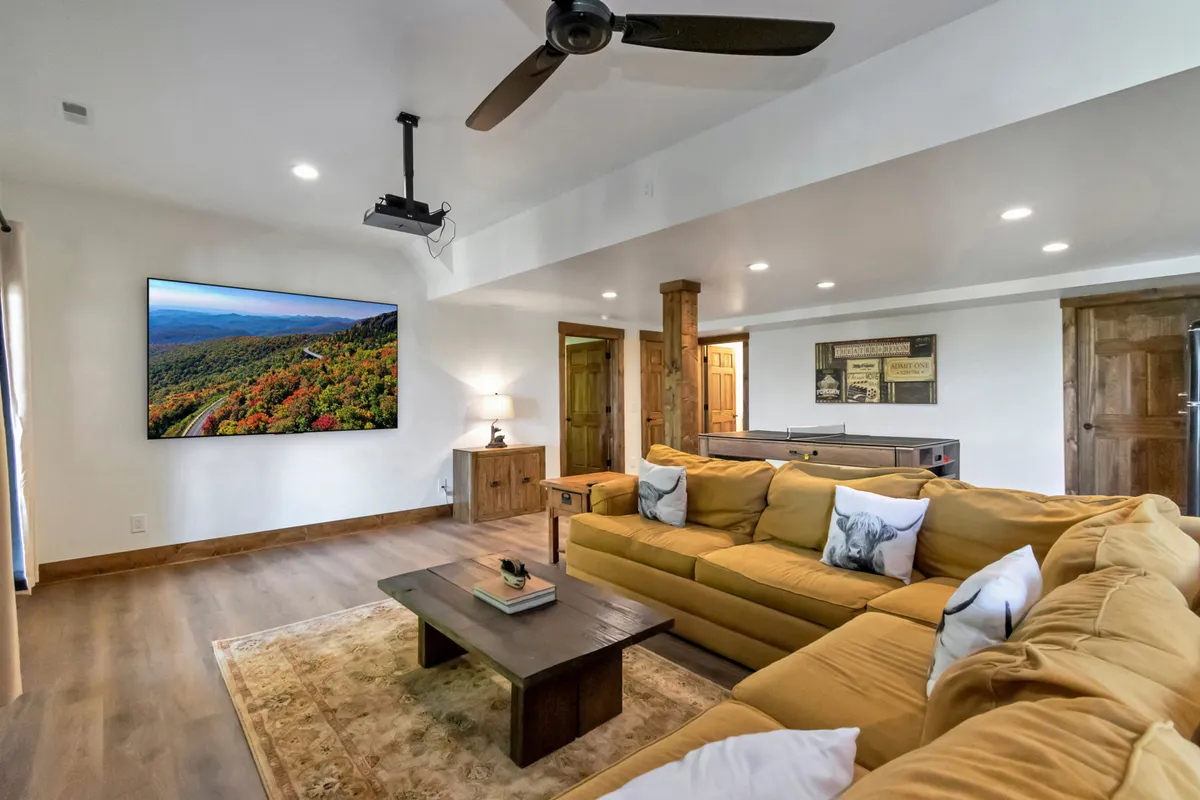This interior shot centers on a large yellow sectional sofa facing a flat-screen TV, with a wooden coffee table and decorative wall art. The TV displays a scenic mountain view, hinting at both entertainment and the natural beauty just outside.