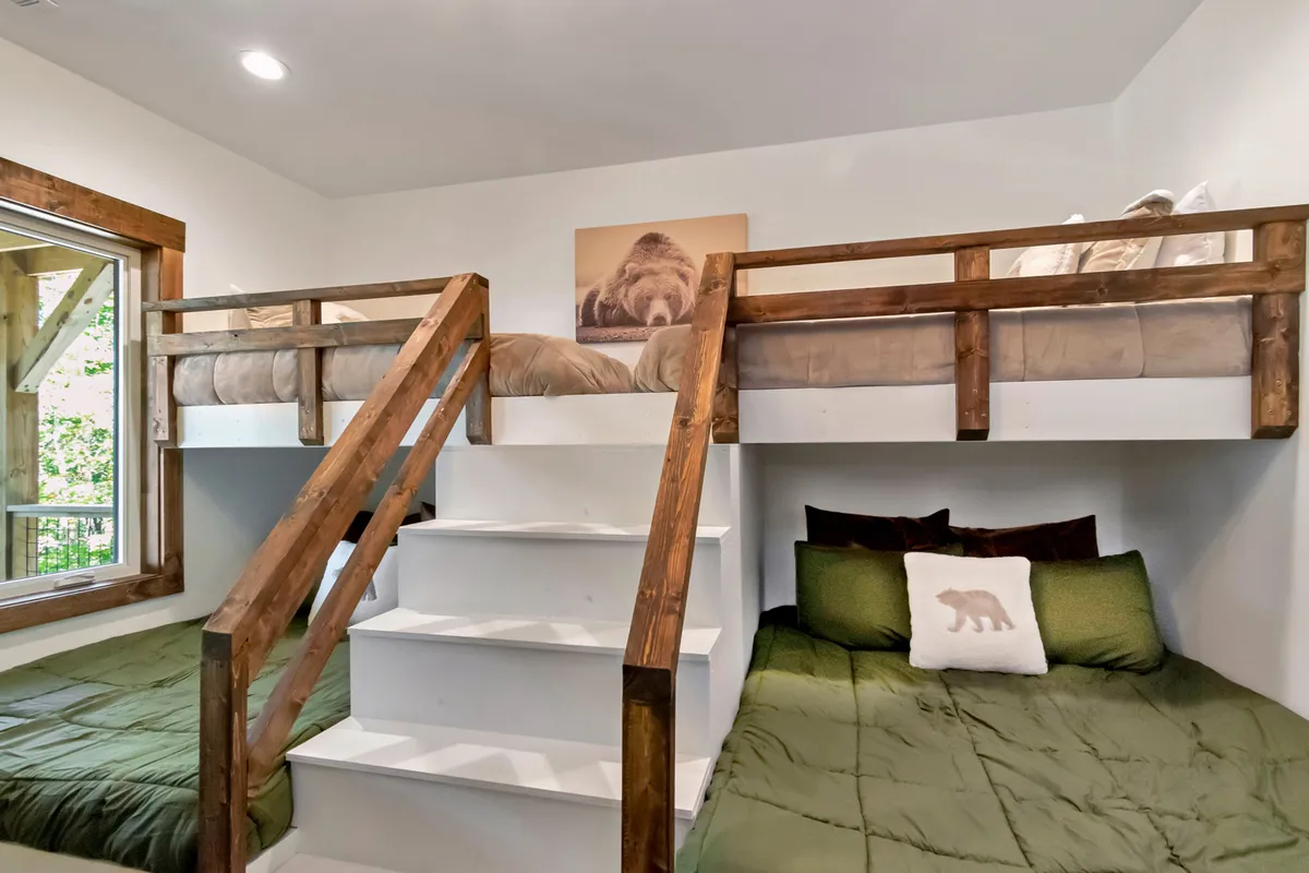 A playful double-bunk room showcases custom wooden bunk beds with green bedding and bear-themed decor. A small staircase leads up to the upper bunks, and the room is set up for both sleep and play, suggesting a perfect space for siblings, cousins, or friends to share.