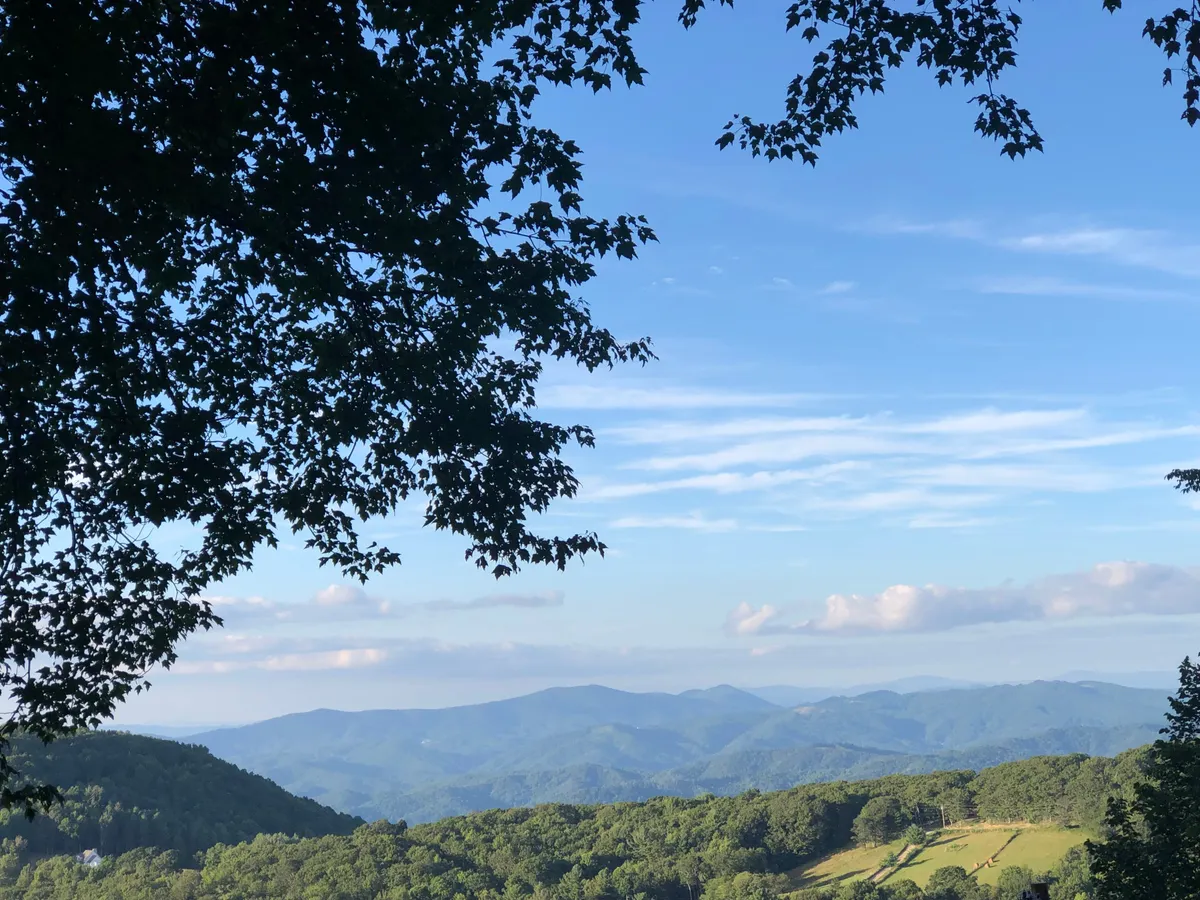 A breathtaking view of a mountain range stretches across the horizon, with lush greenery and a blue sky dotted with clouds. The landscape is open and inviting, capturing the sense of escape and awe that comes with a high-elevation retreat.
