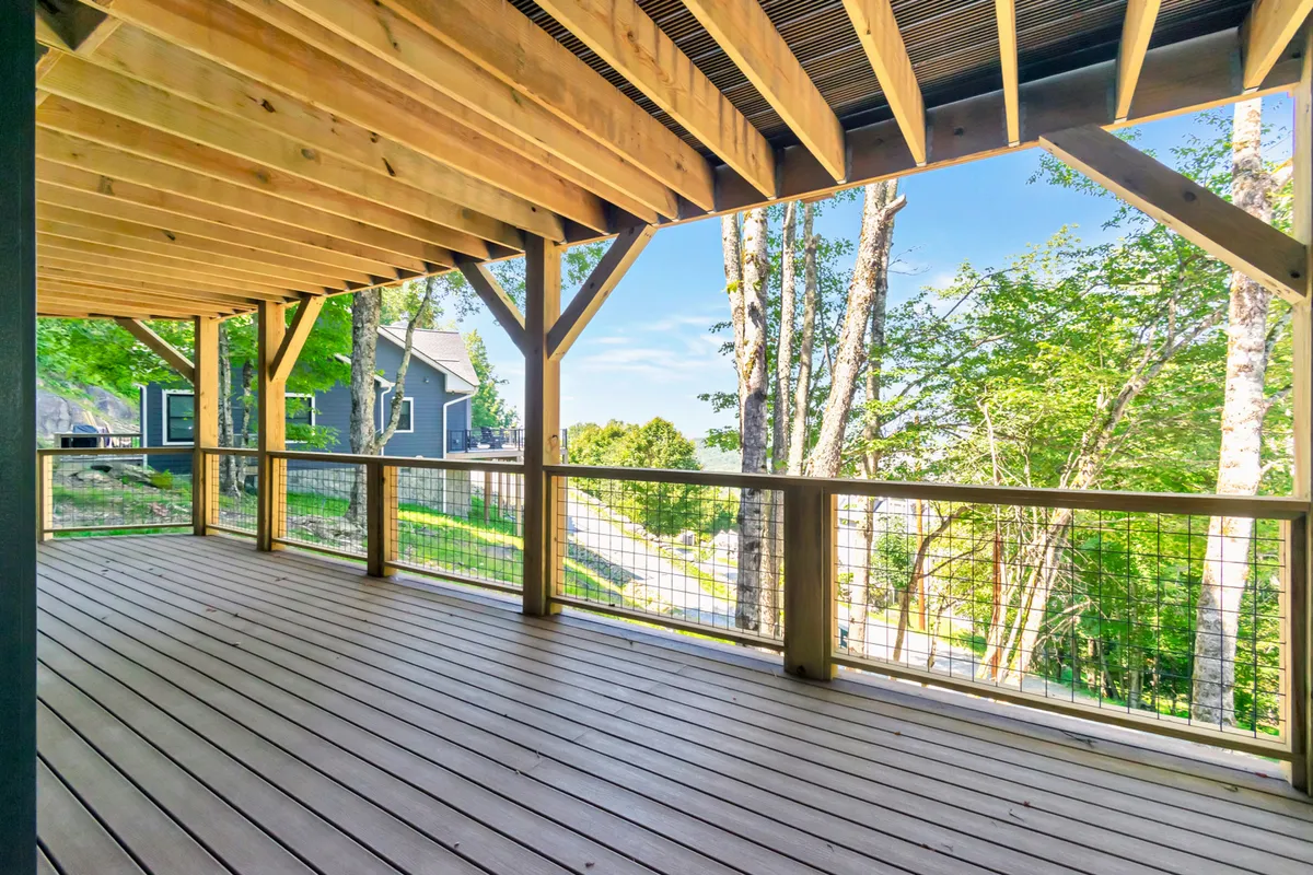 An expansive wooden deck overlooks a tranquil natural setting with trees and neighboring houses in the distance. The deck is bordered by a sturdy wooden railing, suggesting ample space to relax, entertain, or enjoy nature.