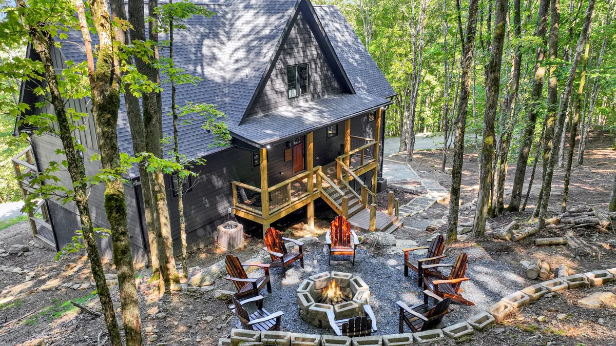 The image showcases a luxurious mountain cabin nestled among lush trees, with a spacious outdoor area featuring a glowing fire pit surrounded by comfortable seating. The cabin’s expansive deck invites relaxation, while the serene natural backdrop suggests privacy and immersion in nature.