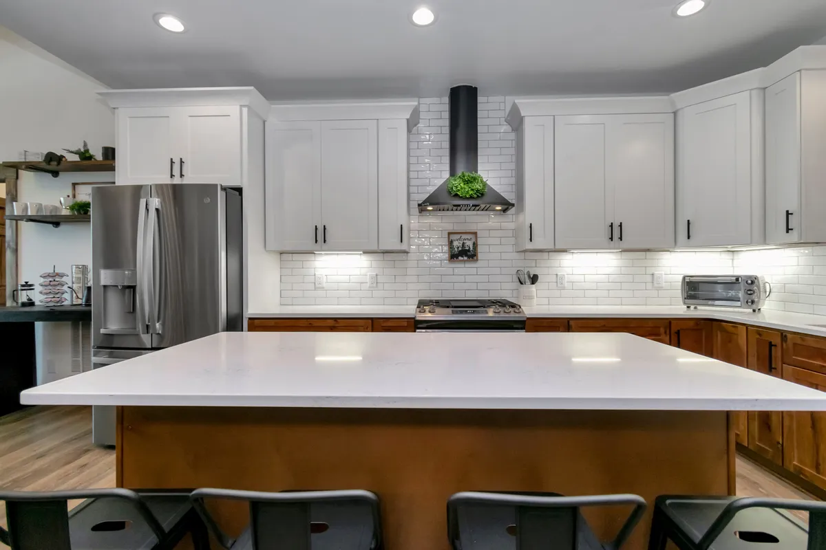 A modern kitchen boasts a generous island, stainless steel appliances, a stylish exhaust hood, and a sleek white subway tile backsplash. The space is designed for both casual breakfasts and evening wine, with inviting touches and ample room for entertaining.