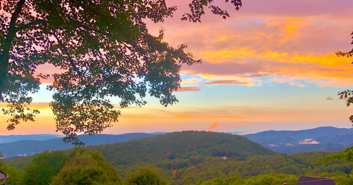 A breathtaking view of a mountain range at sunset, with colorful sky hues and tree branches in the foreground. The wide perspective invites guests to imagine the serenity and natural beauty accessible from the outdoor spaces.